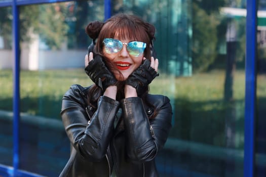 A beautiful girl in a black leather jacket and gloves, wearing blue sunglasses is holding a .headphones on her head in front of the big blue windows.