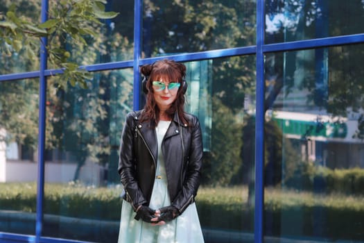 A happy young woman in a blue dress and a black leather jacket, wearing blue glasses against the background of a glass wall, looks at the camera.