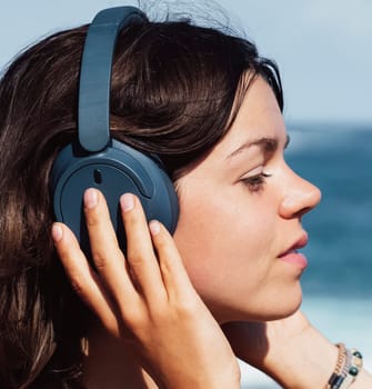 Portrait of a girl with stylish sea blue headphones against the ocean on a sunny day. A young lady walks along the coast and enjoys the sound, listens to music in headphones.