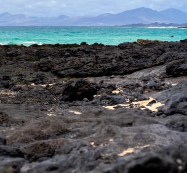 Beach with black volcanic sand and stones on the coast of the Atlantic ocean. A beautiful light blue clean water and mountains on the horizon, a picturesque shore by the sea without people.