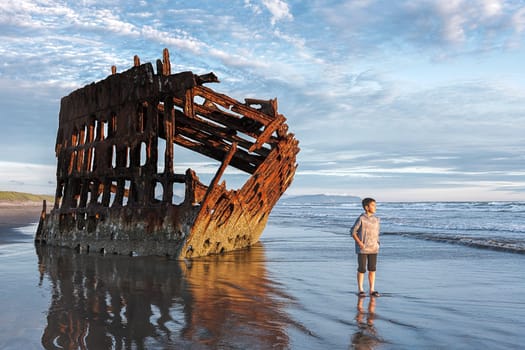 Boy stands by the Peter Iredale shipwreck in the golden sunset light in northwest Oregon.