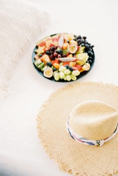 Plate of chopped fruit stands on a white sheet on the bed next to a straw hat. High quality photo