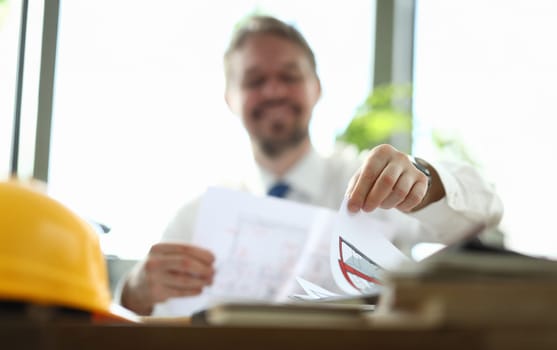 Close-up of businessman holding paper with house plan. Yellow helmet on working table. Smiling person in trendy suit. Modern renovation agency and perfect service concept