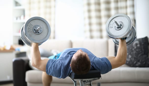 Close-up of strong and fit male lifting heavy dumbbells indoors. Sport and active lifestyle. Person laying on back and training for health and body. Quarantine period concept