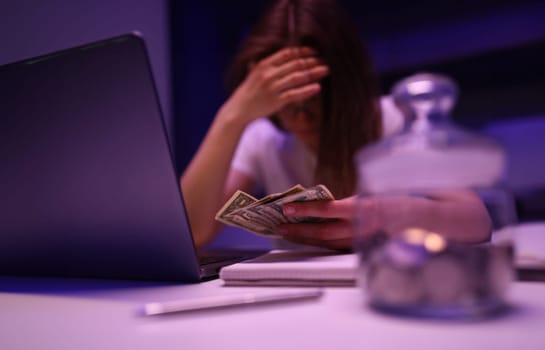 Close-up of depressed woman calculating family financial situation. Person close face with hand and holding banknotes. Glass container with coins. Money and investment concept