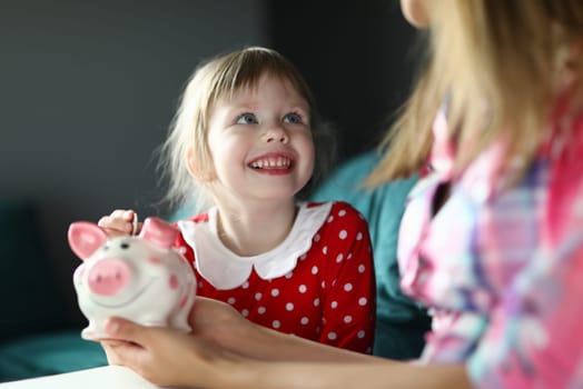 Portrait of smiling cheerful kid putting coin in piggy bank and looking at mother. Happy joyful child wearing red dress in dots. Investment and finance concept