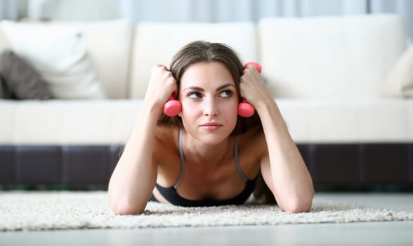 Portrait of tired young woman taking pause in train. Pretty female laying on fluffy carpet at home holding pink dumbbells. Sport and hobby. Indoors workout on quarantine concept