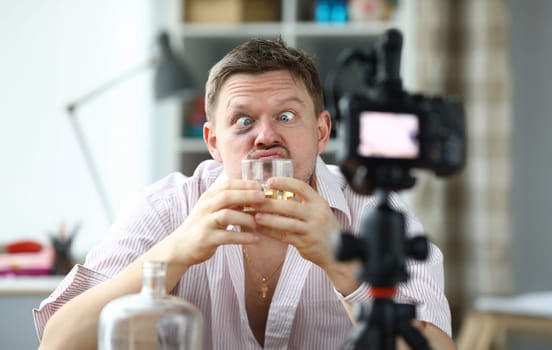 Portrait of drunk male filming on camera smelling glass of whiskey. Middle-aged man making funny face and look messy. Tasting of alcoholic drink on videocamera. Blogger concept