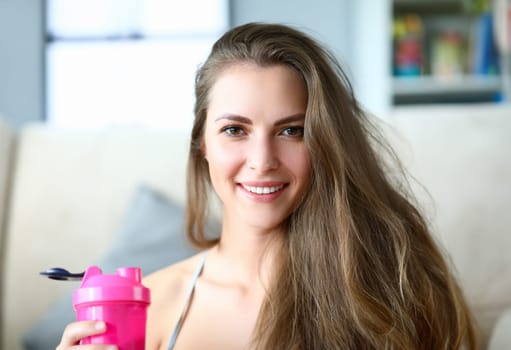 Portrait of cute young woman holding pink bottle with water after workout. Happy smiling person posing on camera. Tired after training at home. Sport and active lifestyle concept