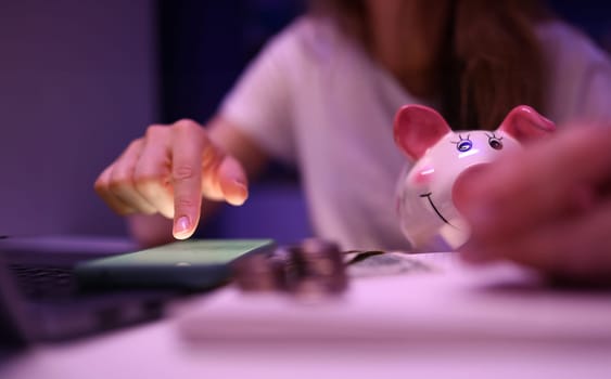 Close-up of person accounting money on mobile phone. Pink piggy bank banknotes and coins on table. Detailed shooting. Family budget. Finance and investment concept