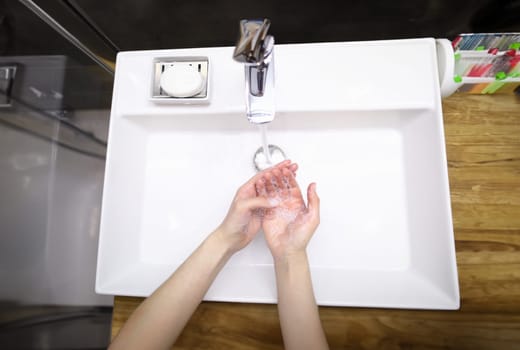 Top view of person washing hands with soap under water. Prevention infection spread. Clean white modern faucet. Silver crane. Bathroom and personal hygiene concept