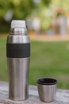 Stainless steel cup, mug with thermos stands on a wooden table in the forest on a green natural background. Thermos and mug with hot drink standing on wet wooden table after rain outdoors, close-up. Steam rises from thermo mug