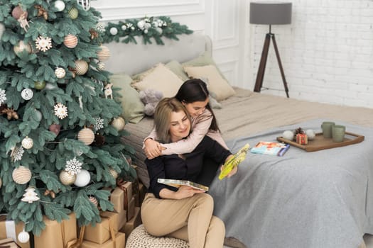 Beautiful young mother with long hair is reading a photo book with her little daughter. christmas home decor. warm tinted photo