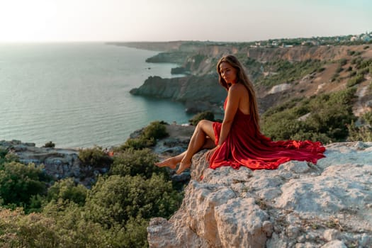 Woman sunset sea red dress, side view a happy beautiful sensual woman in a red long dress posing on a rock high above the sea on sunset