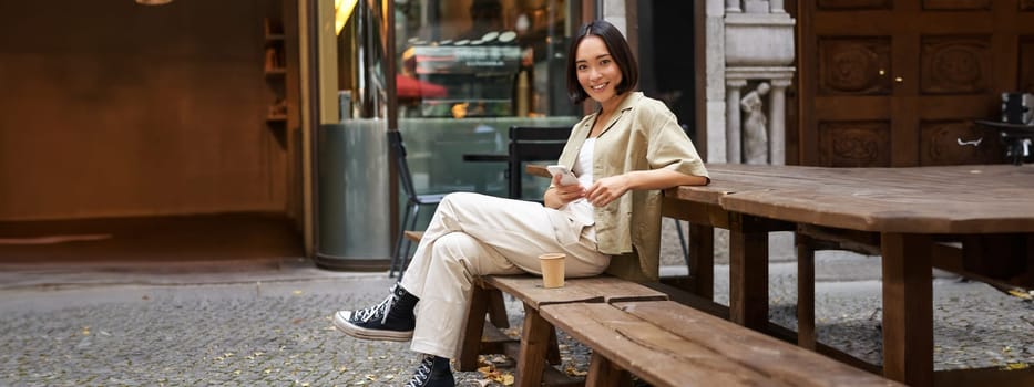 Portrait of stylish young korean woman sits in cafe, holds smartphone, smiles, enjoys coffee outdoors.