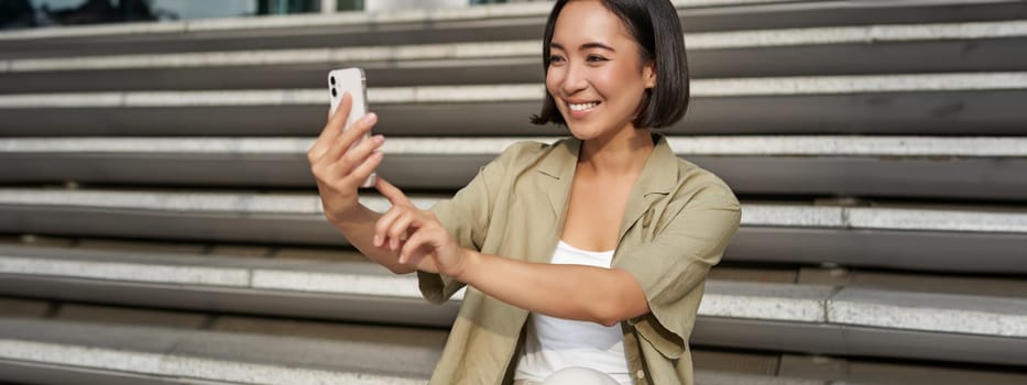 Portrait of asian beautiful girl takes selfie on smartphone. Young korean woman sits on stairs outdoors and makes photos on mobile phone.