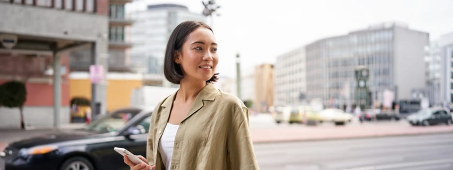 Young woman walking down the city street, turn behind, using map app navigator on smartphone, holding mobile phone.