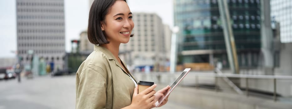 Beautiful girl standing in city square with tablet, reading and drinking takeaway coffee.