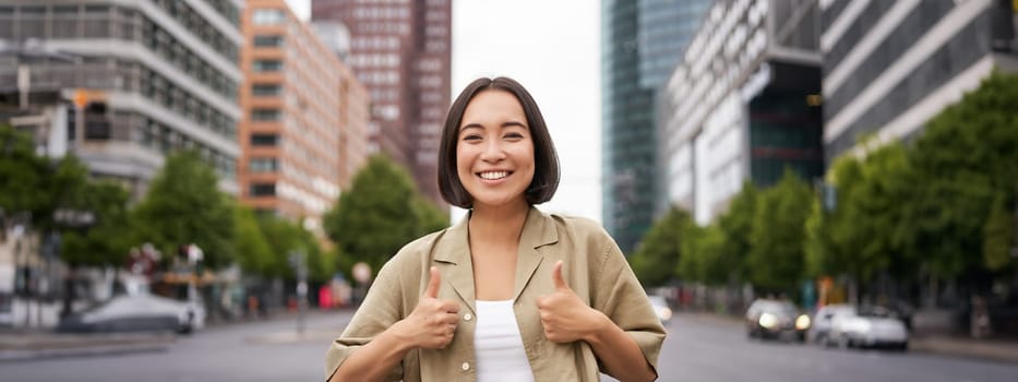 Enthusiastic city girl, shows thumbs up in approval, looking upbeat, say yes, approves and agrees, stands on street.