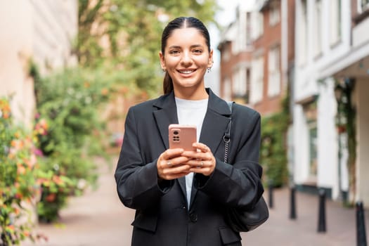 Sunny portrait of a newly entrepreneur young woman in her 25s, black suit, holding a phone smiling, looking in camera.