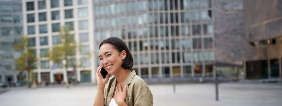 Vertical shot of smiling stylish asian girl, having a telephone conversation and walking on street. Young woman talking on mobile phone.