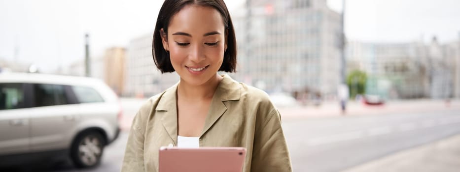 Portrait of asian girl smiles, reads on tablet, uses device while stands on city street.