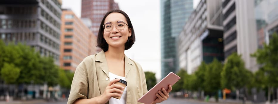 Portrait of happy young woman in glasses, standing on street with cup of coffee and tablet.