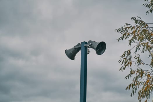 Tall metal column with two gray loudspeakers against cloudy sky. Hazard warning system. Providing security in the city, notification of emergencies. Copy space for text.