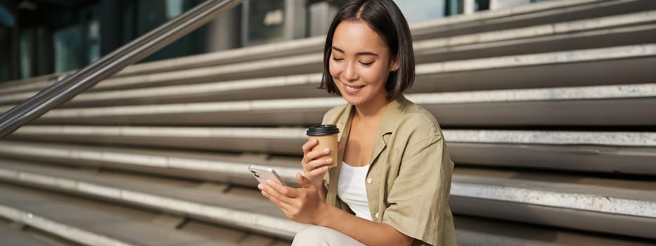 Happy urban girl drinks her takeaway coffee and scrolls feed on smartphone. Asian woman sits on stairs with tea and holding mobile phone.