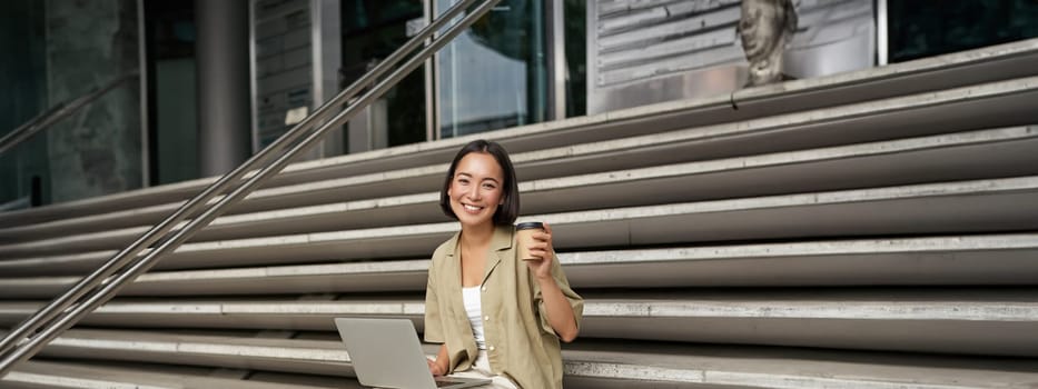 Vertical shot of smiling girl student, asian woman sits on stairs of university campus and drinks coffee, does her homework on laptop.