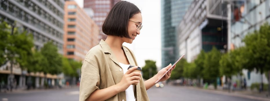 Happy woman exploring the city. Young korean girl holds tablet, drinks coffee and walks along street with big smile on her face.