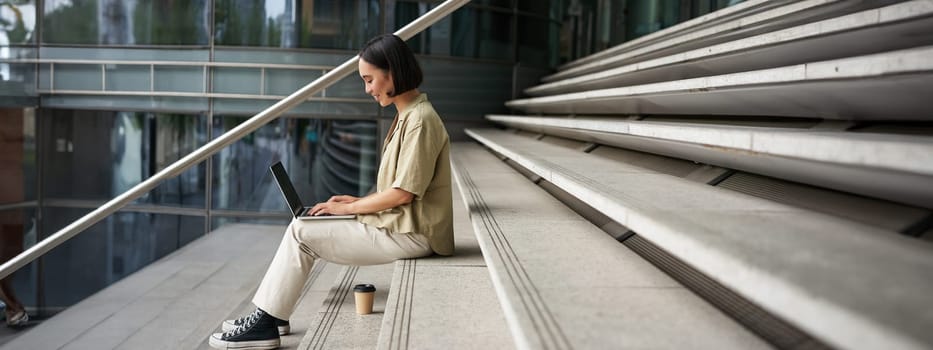 Profile portrait of young asian woman with laptop, girl student sits on stairs outside building and types on computer, drinks takeaway coffee.