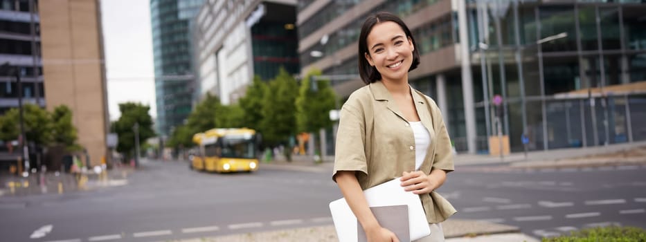 Beautiful asian girl smiles as commutes to work, stands on street with laptop and notebook.