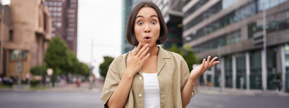 Modern asian girl standing on street with surprised face, looking impressed and amazed, hear awesome news.