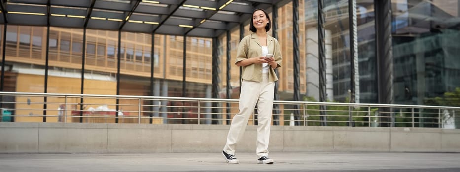 Portrait of young asian woman, student with smartphone, standing on street near glass building, waiting for someone.