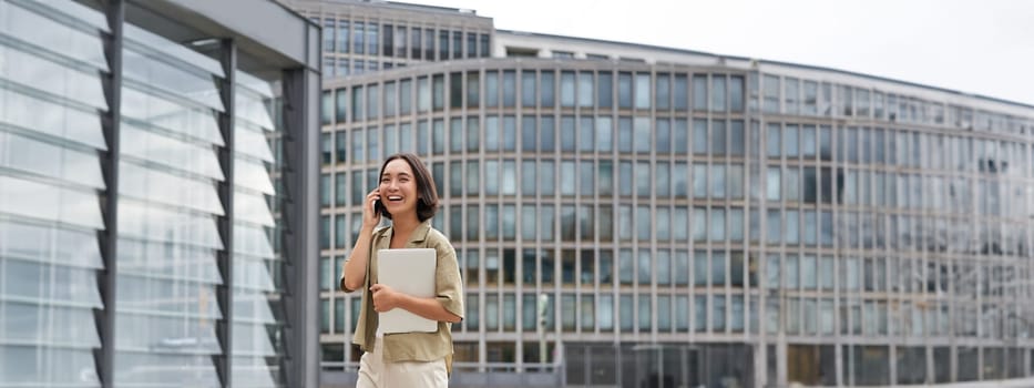 Stylish urban girl walking on street, talking on mobile phone and carrying laptop. Young woman making a telephone call while going somewhere.