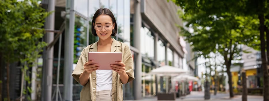 Outdoor shot of young korean woman stands on street with laptop, wears glasses, reads, smiles happily.