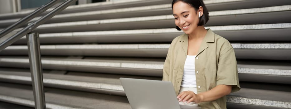 Remote worker. Smiling asian girl sits outdoors on street with laptop. Happy young woman working on computer remotely, listen music in wireless headphones.