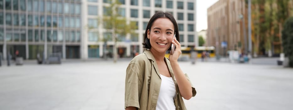 Modern young asian girl talks on mobile phone, uses telephone on city street. Woman smiling while calling someone on smartphone.