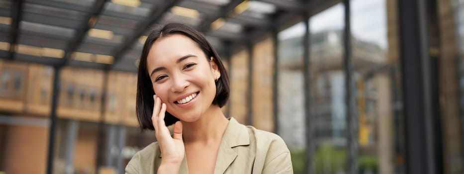 Close up portrait of asian girl with happy face, smiling and laughing, touching her skin, standing on street on sunny day.