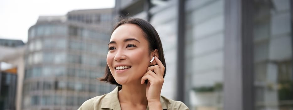 Vertical shot of smiling asian woman in wireless headphones, enjoys listening to music in earphones, holds mobile phone.