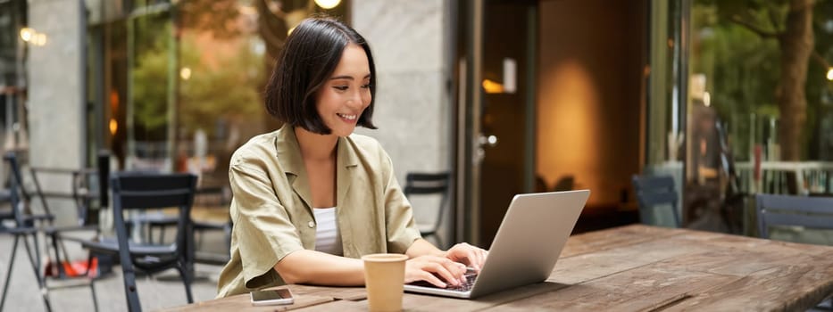 Young asian woman, digital nomad working remotely from a cafe, drinking coffee and using laptop, smiling.