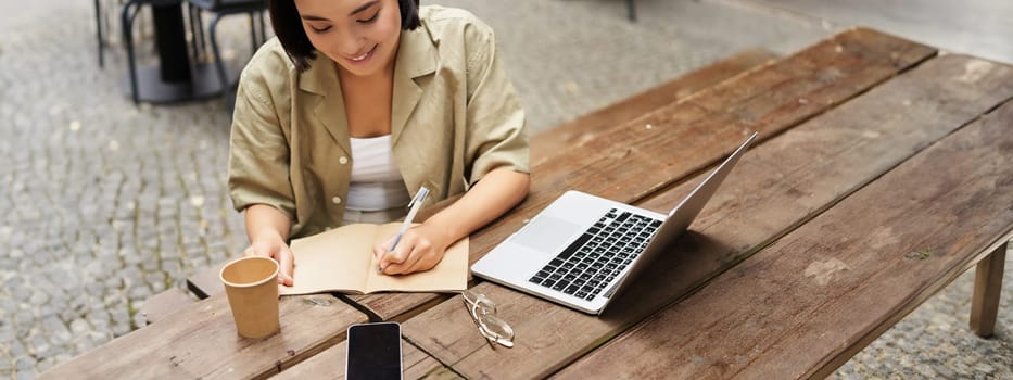 Portrait of young woman studying online, sitting with laptop, writing down, making notes and looking at computer screen, sitting in cafe outdoors.