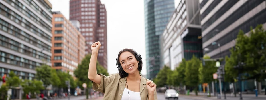 Portrait of smiling asian woman dancing, triumphing, feeling happy while listening music in city, posing on street near skyscrappers.