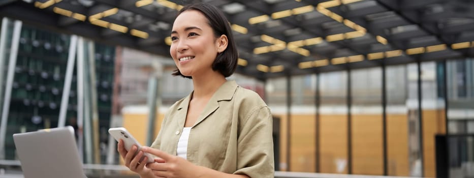 Technology and people. Smiling asian girl with mobile phone, using telephone and walking in city in daylight.