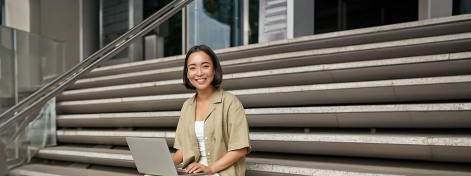 Vertical shot of asian girl sits with laptop, drinks coffee on university stairs. Young woman, student does her homework outdoors.