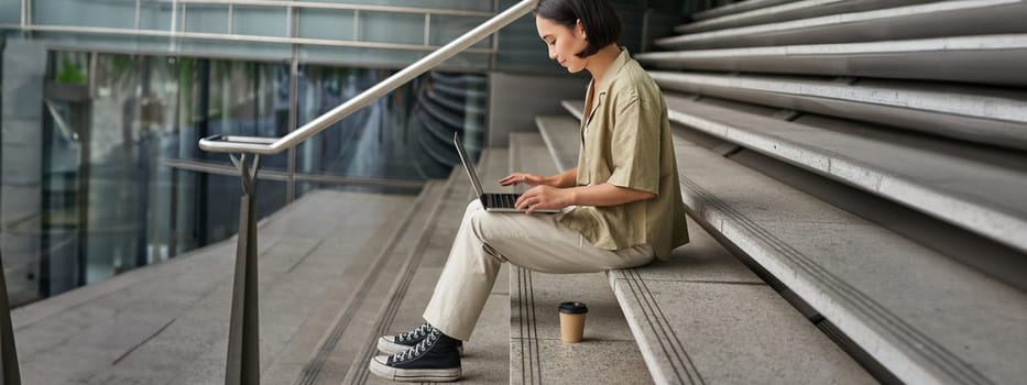 Profile portrait of young asian woman with laptop, girl student sits on stairs outside building and types on computer, drinks takeaway coffee.