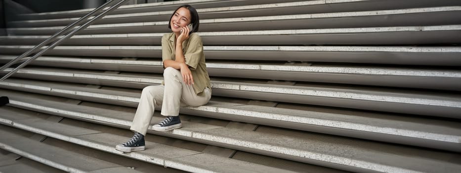 Portrait of asian student girl on stairs talks on mobile phone, smiles at camera, sits outside building.