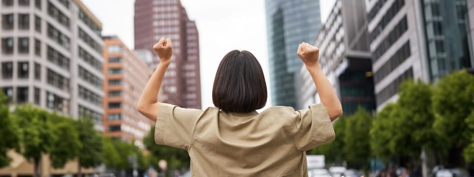 Excitement. Happy girl rear view, raising her hands up and looking at big city skyscrappers, feeling confident, cheering and triumphing.