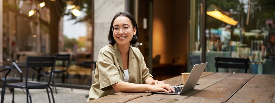 Portrait of asian girl works in cafe, uses laptop and sits outdoors on street. Digital nomad and online learning concept.